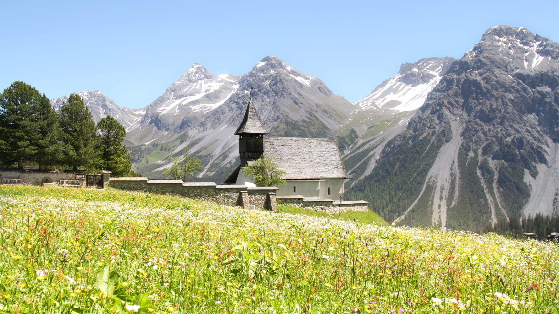 Stone church with steeple in wildflower meadow with snow-capped mountain peaks under blue sky in background.