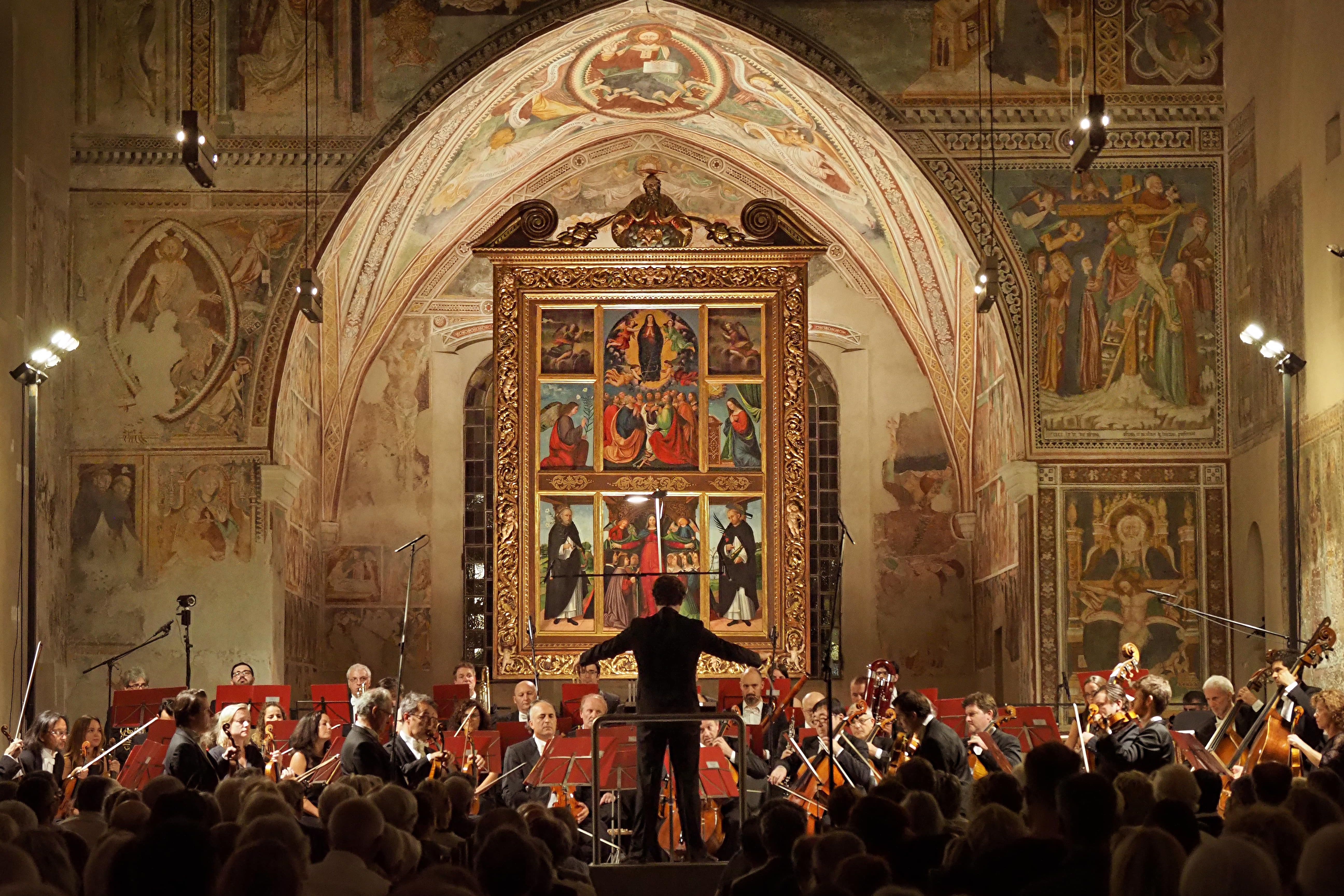 Orchestra performing in historic church with ornate frescoed arch and gilded religious altarpiece, conductor facing musicians.