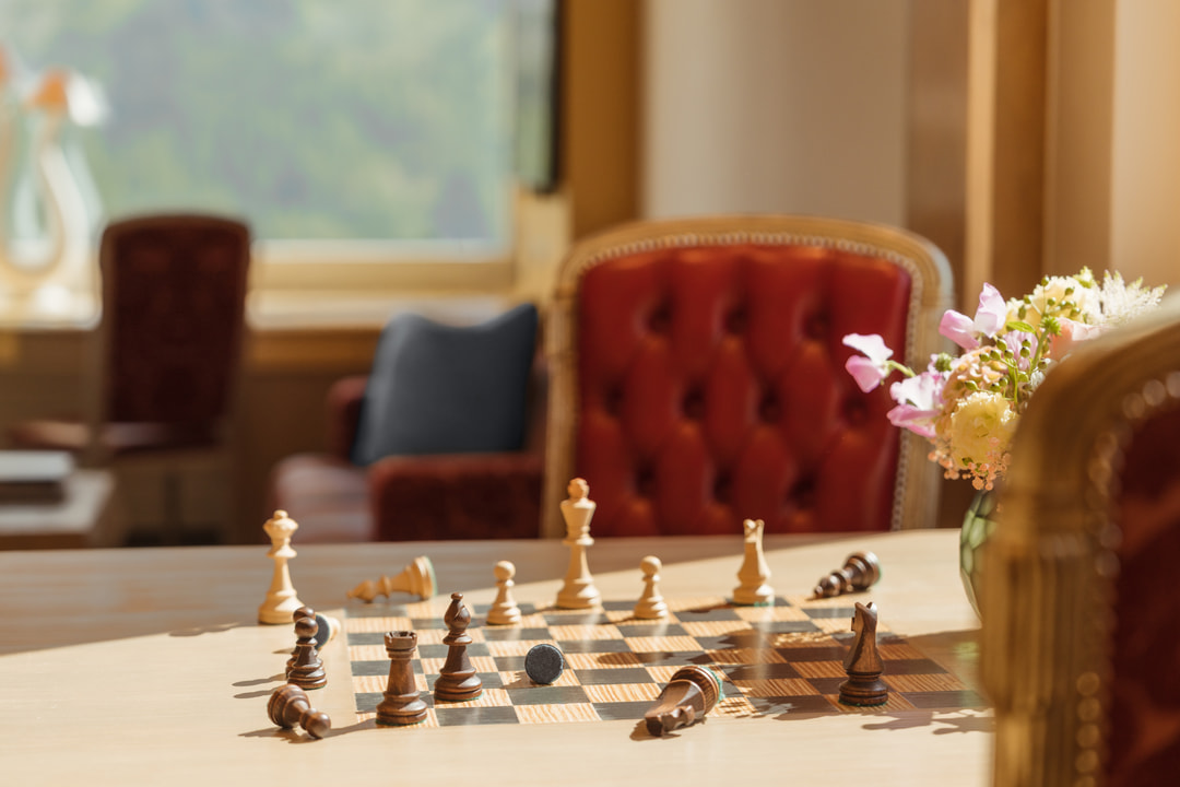 Close-up of a chessboard on a wooden table in a bright hotel living area, with red upholstered armchairs and a floral arrangement softly blurred in the background.