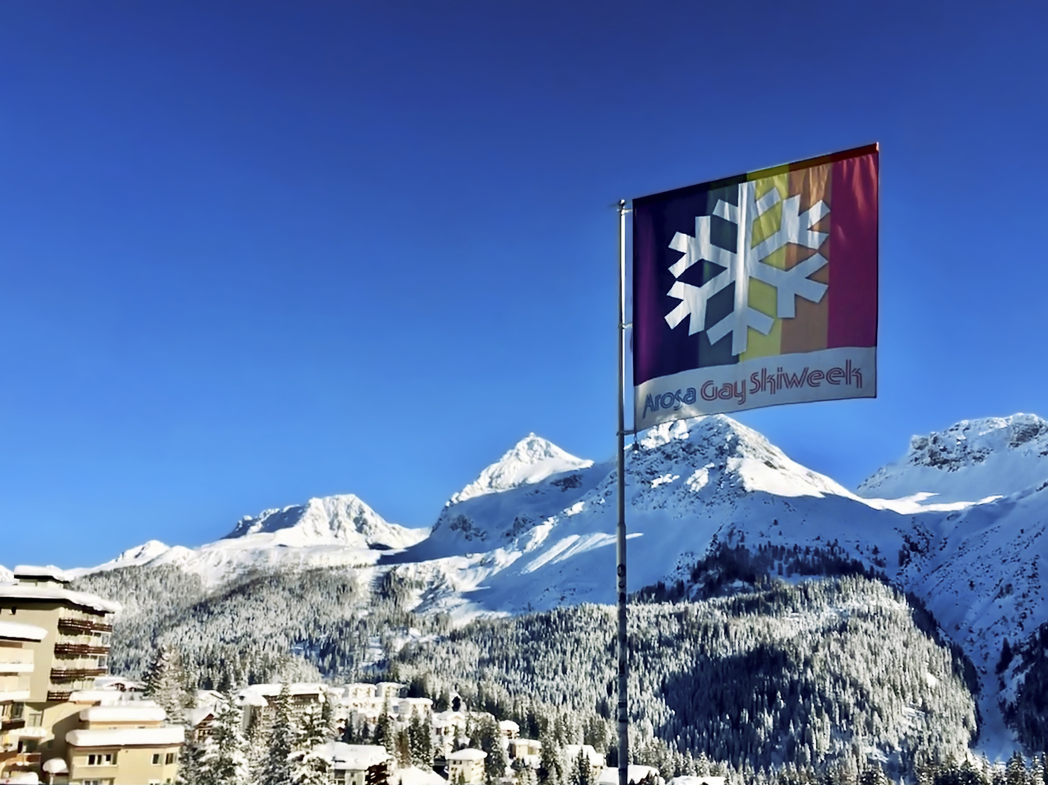 Arosa Gay Ski Week flag with snowflake logo flying against snow-covered mountains and blue sky in Swiss Alps resort.