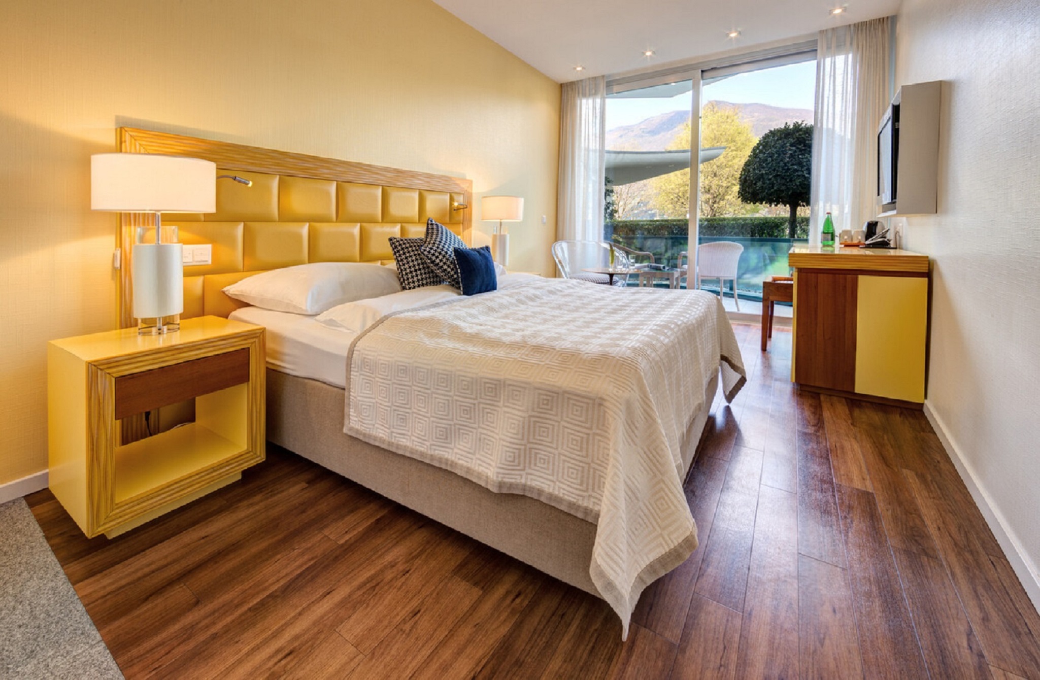 Sunny hotel room with yellow upholstered bed, matching nightstands, wooden flooring, cozy seating area, flat-screen TV, and balcony with orange awning.