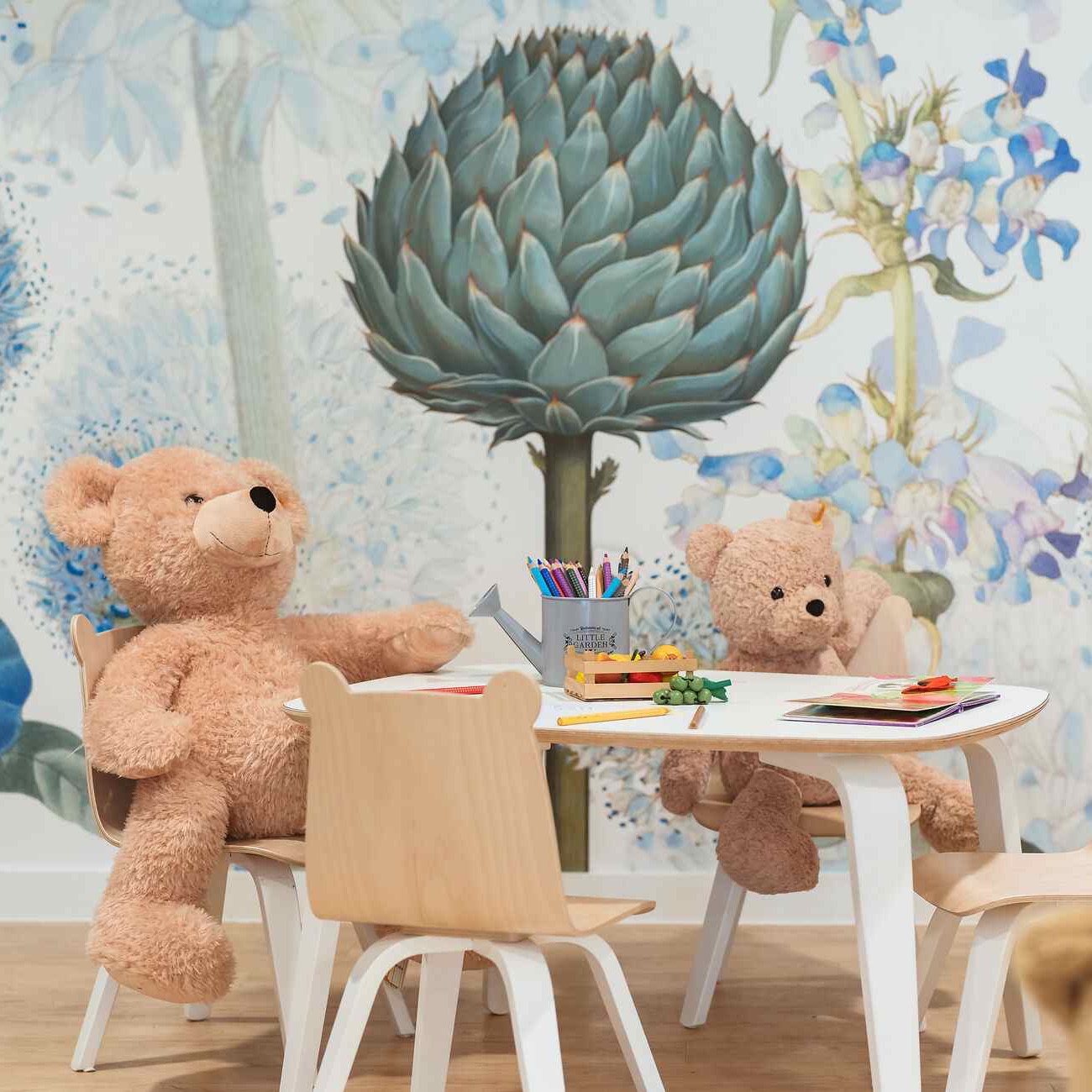 Two plush teddy bears seated at a small table with art supplies against a floral wallpaper backdrop with blue flowers.