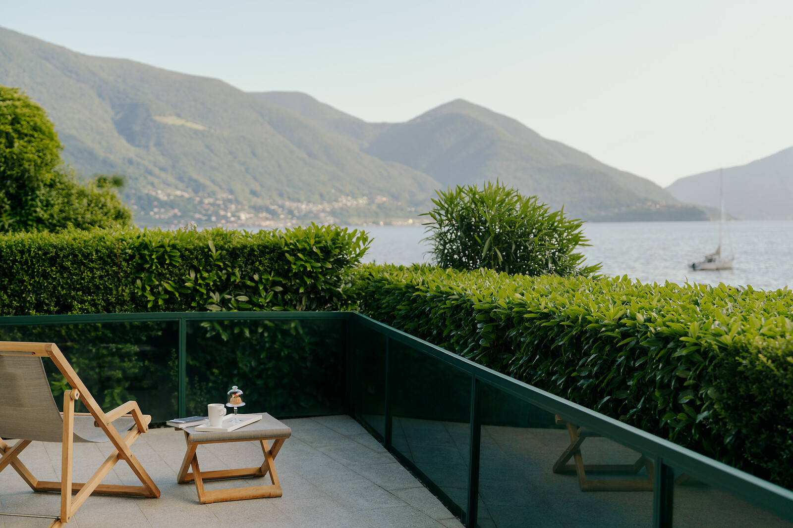 Terrasse mit Liegestuhl und Blick auf einen See mit Bergen im Hintergrund, grüne Hecken und Segelboot auf dem Wasser.