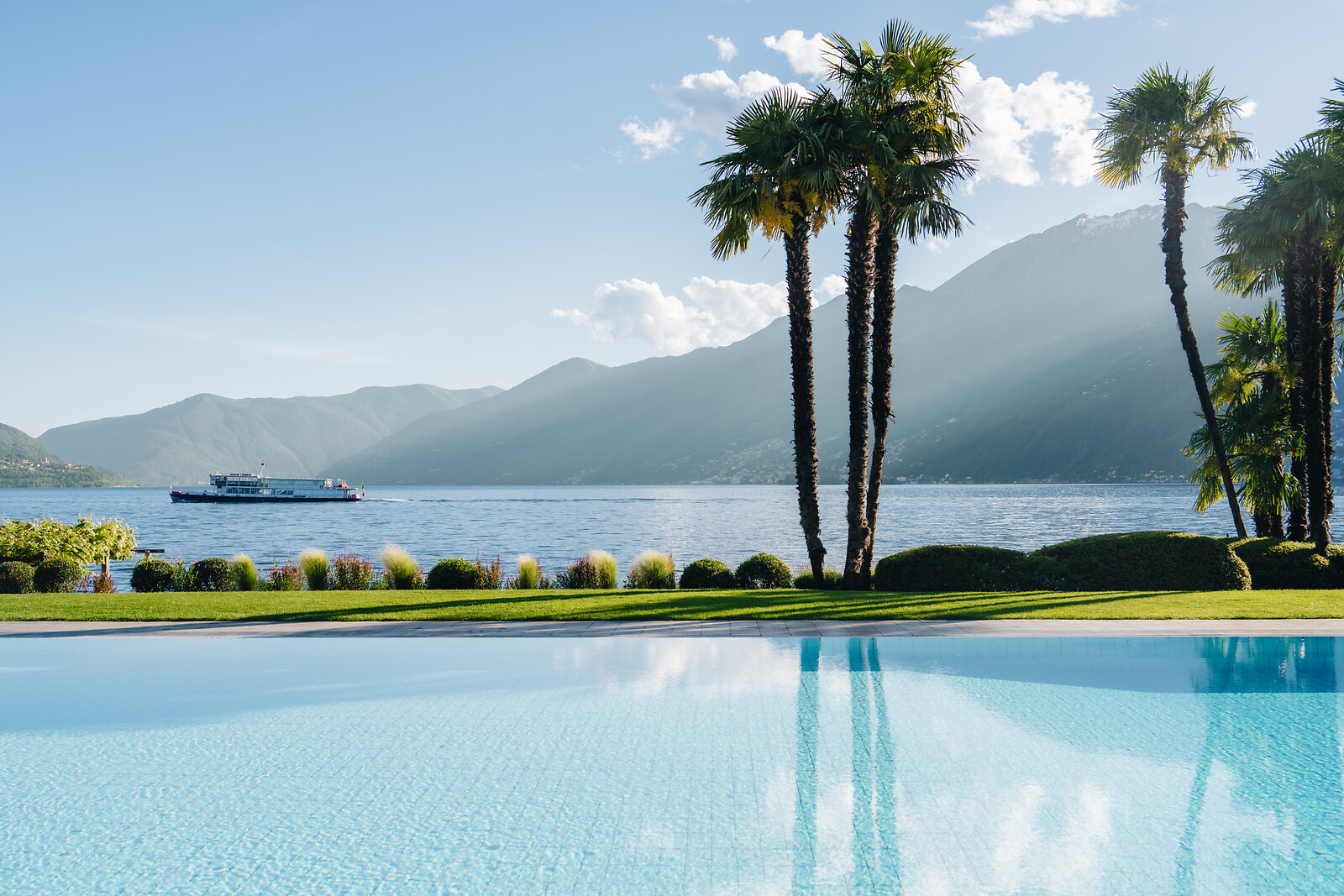Pool mit Palmen, See, Fähre und Bergen im Hintergrund unter blauem Himmel mit weissen Wolken.
