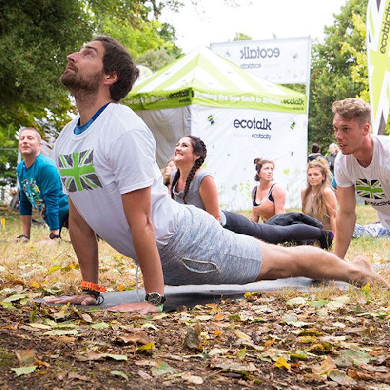 People doing yoga on a leafy piece of ground wearing white t-shirts with the green union jack on them