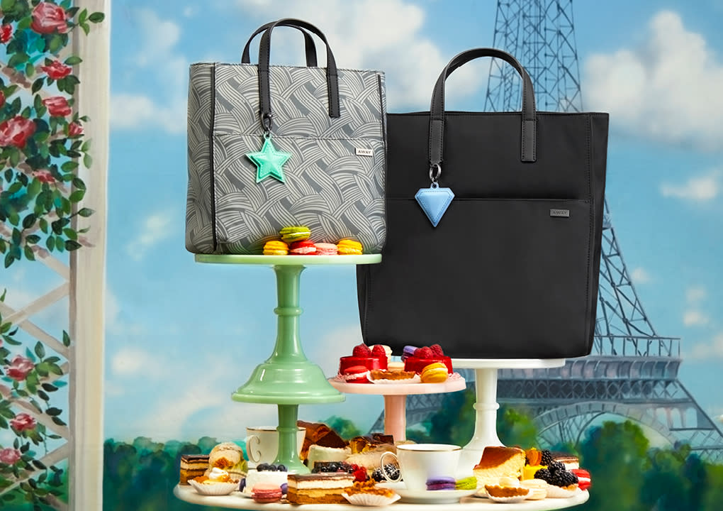 Away Tote shoulder bags in grey and black displayed on food servers with dessert against a background of the Eiffel tower.