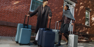 Two travelers effortless rolling 6 pieces of Away luggage as they walk along a sidewalk in front of a brick building, smiling at each other.