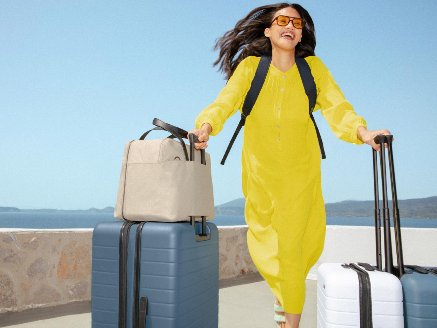 A woman in a yellow dress carrying several Away luggage pieces, including a Large Flex suitcase, an Everywhere Bag, and two carry on suitcases in white and coast