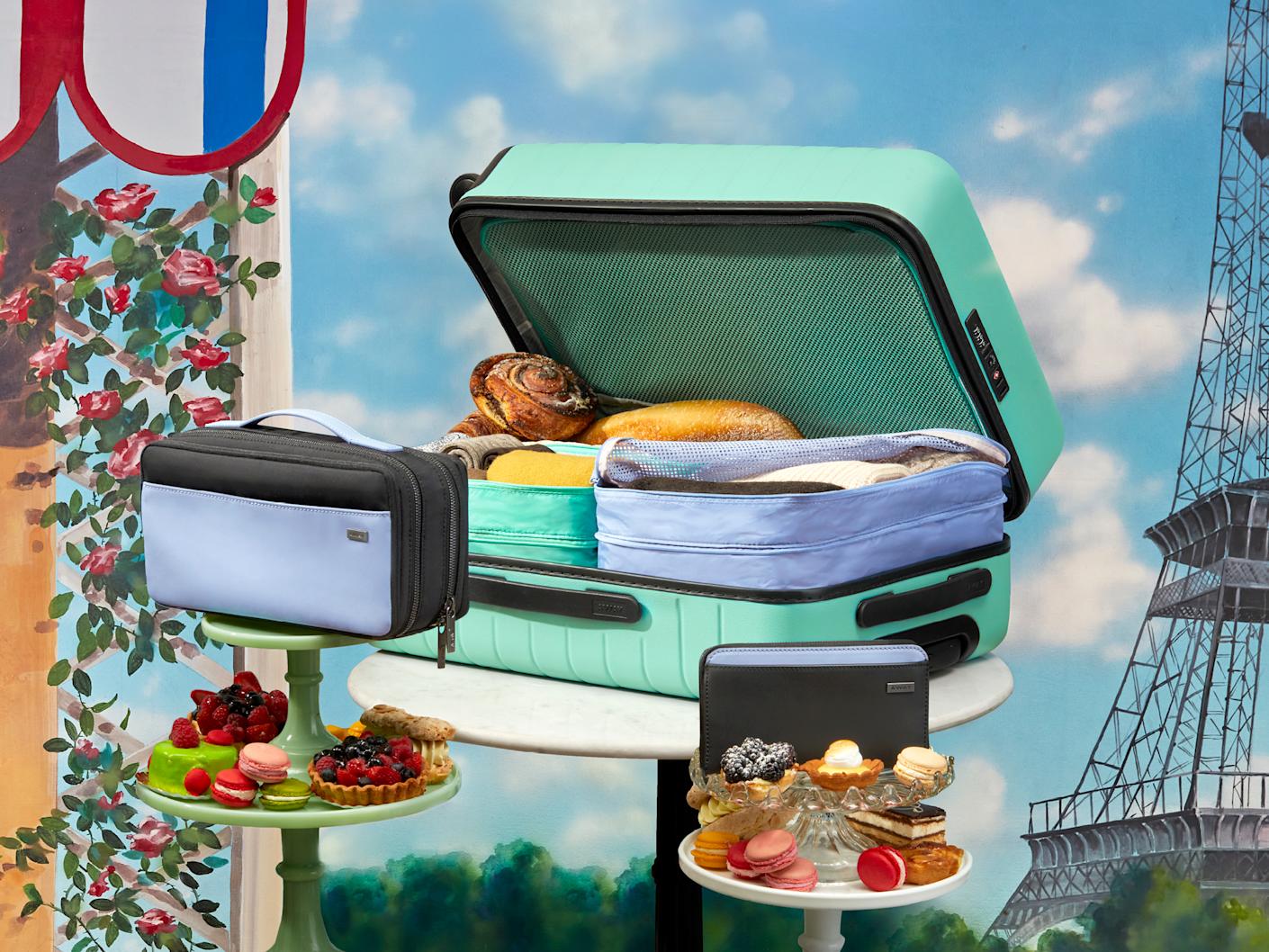A sea green suitcase packed with picnic food, Away travel organizers displayed on platters with dessert with the Eiffel tower in the background.