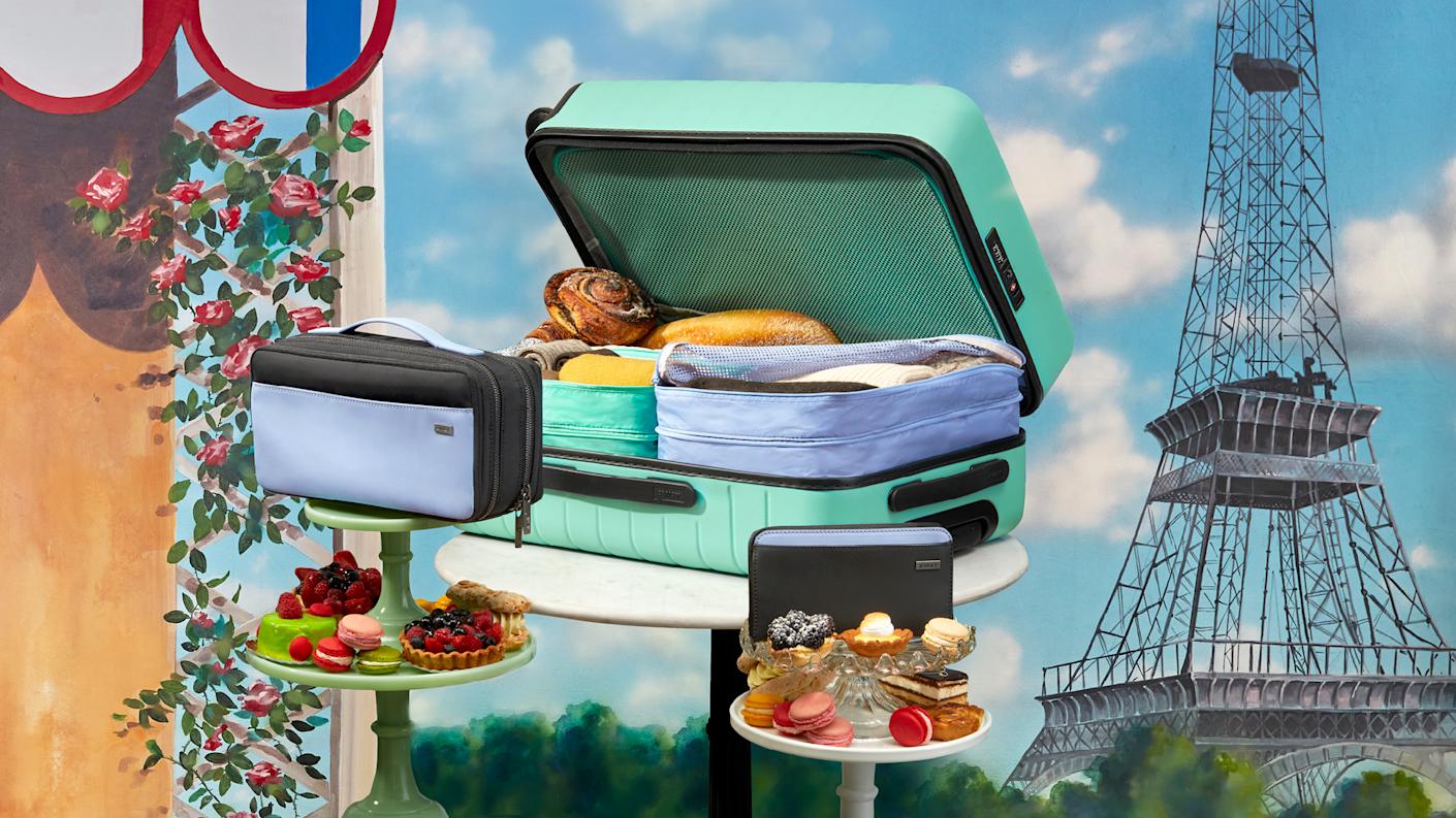 A sea green suitcase packed with picnic food, Away travel organizers displayed on platters with dessert with the Eiffel tower in the background.