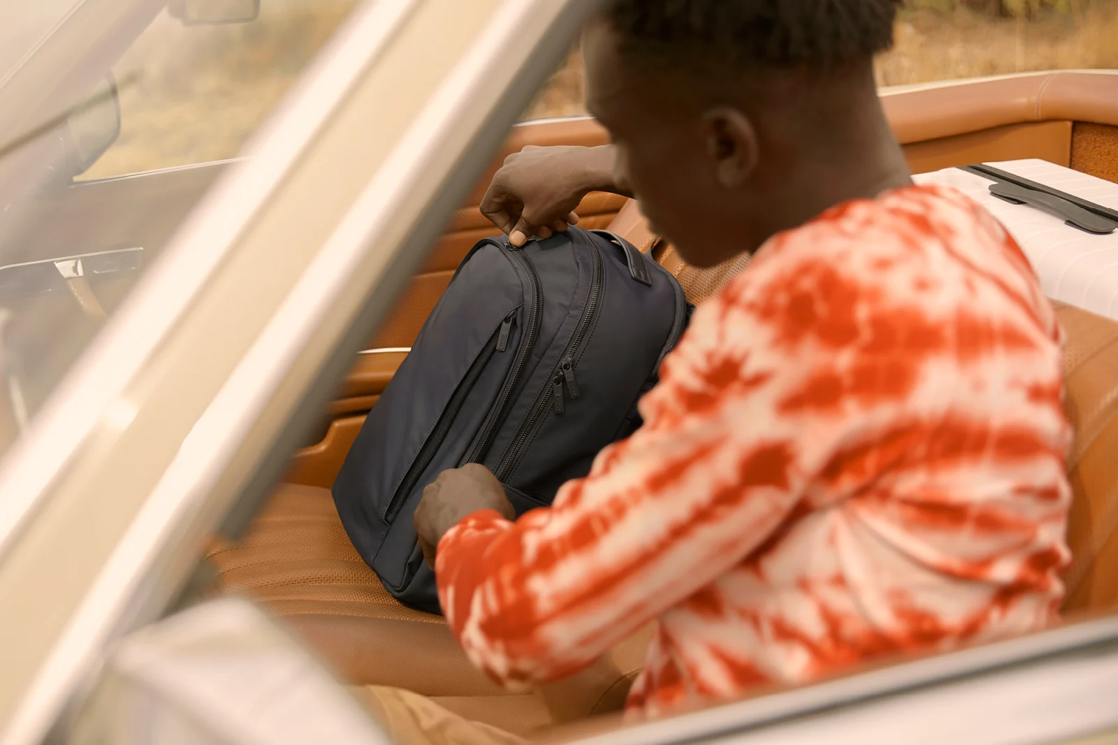 A man with a tie and dye red and white shirt sitting in a car taking something out of his navy blue backpack.