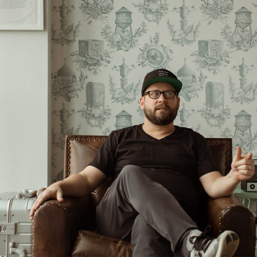 Dallas Taylor, podcast host in a black t-shirt sitting on a sofa, legs crossed.