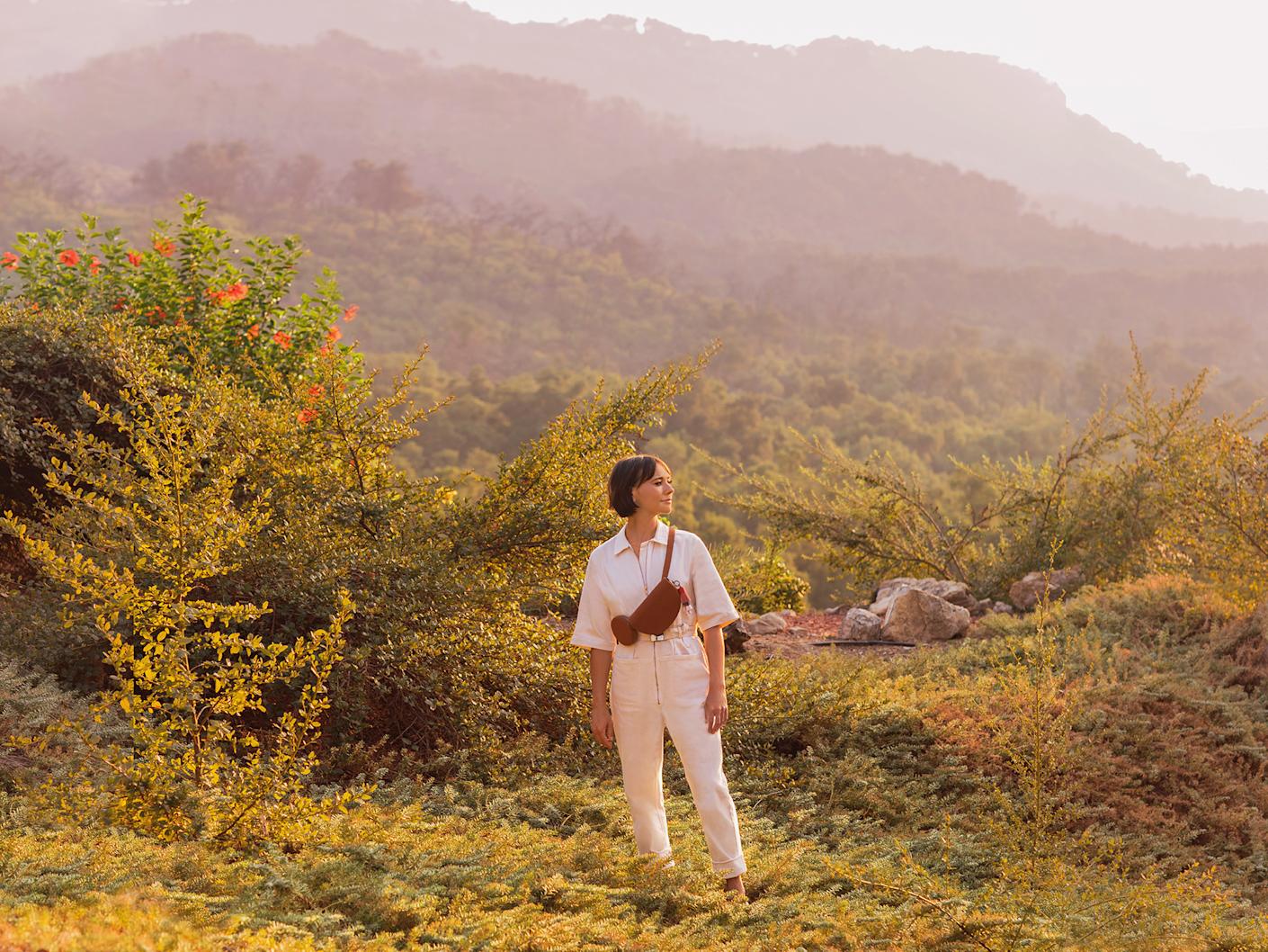 A woman standing in a mountain setting with a crossbody sling bag in brown.