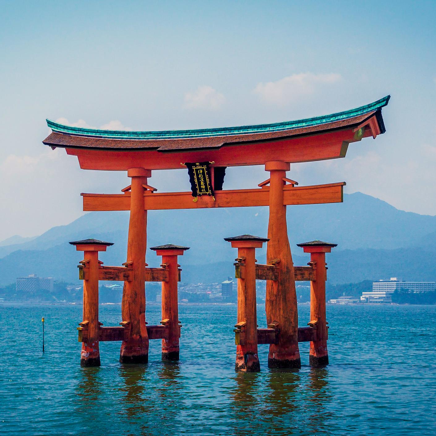 Japanese shinto Itsukushima Shrine in orange partially submerged in water set amidst a mountain backdrop.