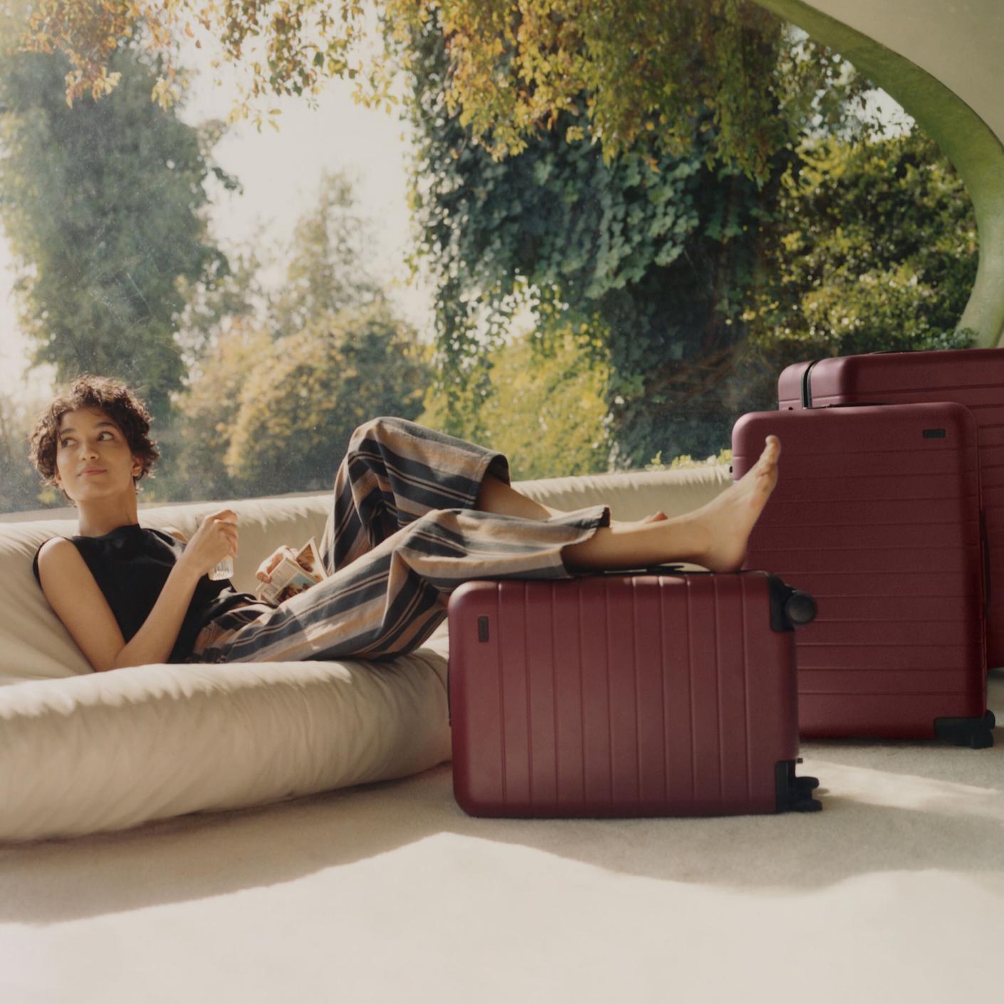 A woman lounging on a sofa ready to travel with her carry-on and checked Away luggage in maroon, at her feet.