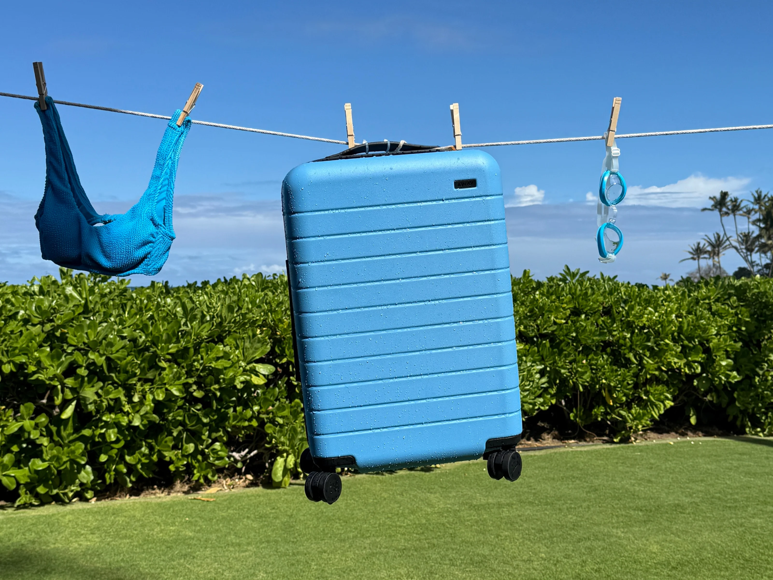 Away Bigger Carry-On in limited edition Splash Blue hanging from a laundry line with other warm weather escape essentials