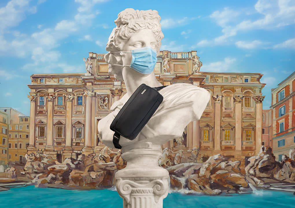 A statue wearing a mask with a black Away sling bag cross bodied against its torso with the Trevi Fountain in the background.