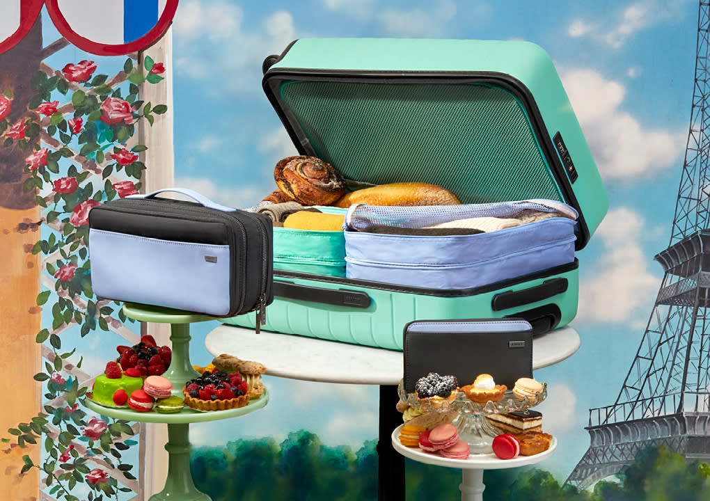 A sea green suitcase packed with picnic food, Away travel organizers displayed on platters with dessert with the Eiffel tower in the background.