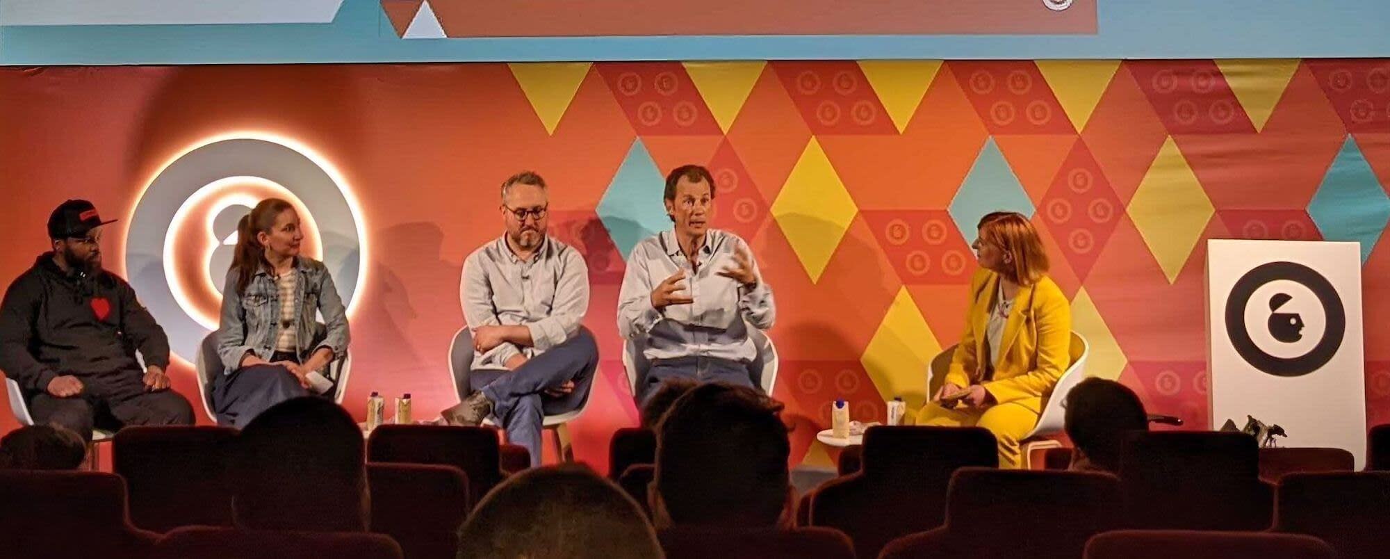 Five people sit on a stage, with brightly-colored lights and shapes behind them.