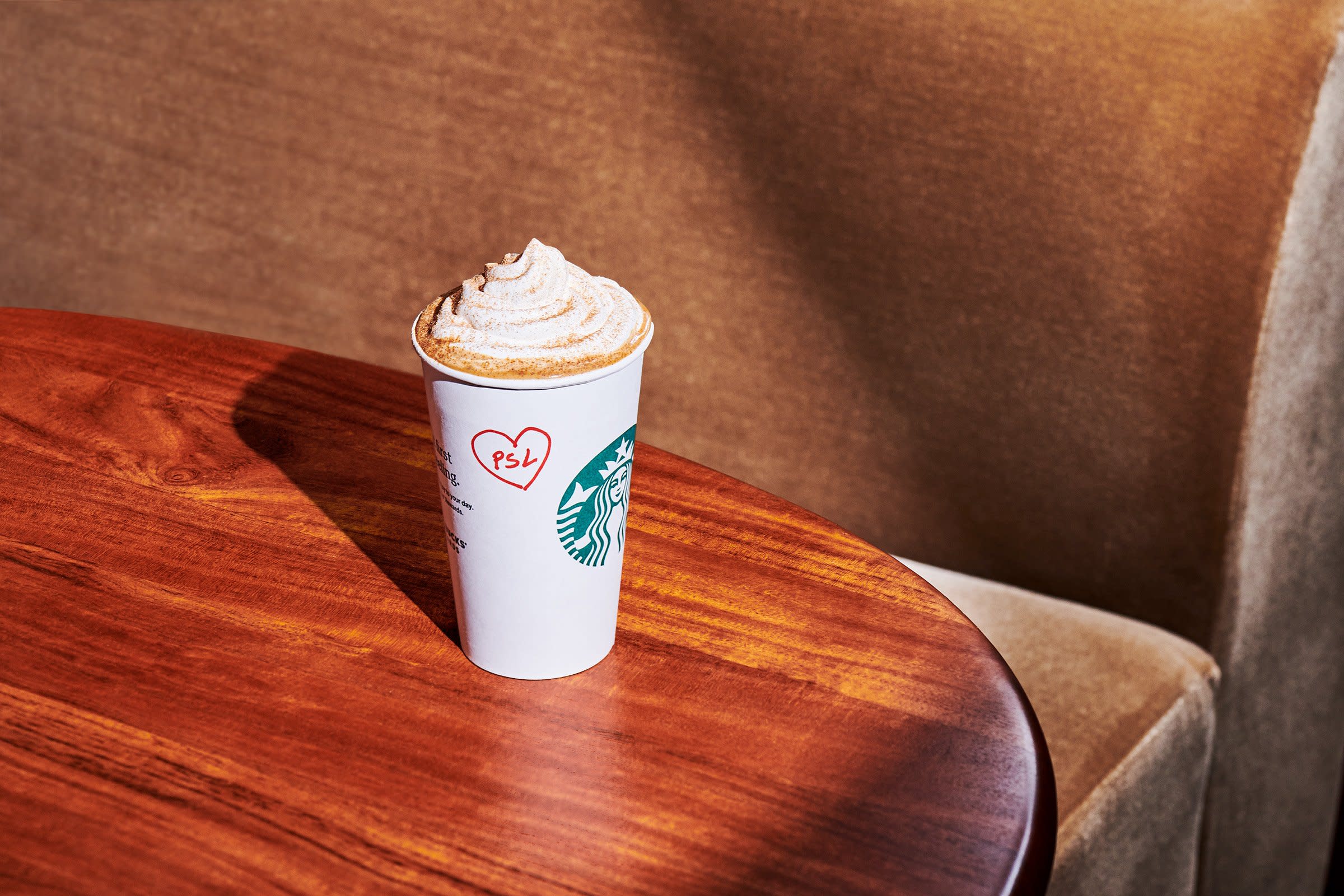 Starbucks' Pumpkin Spice Latte PSL on a wooden table topped with whipped cream with a red heart around the letters "PSL" written on the cup 