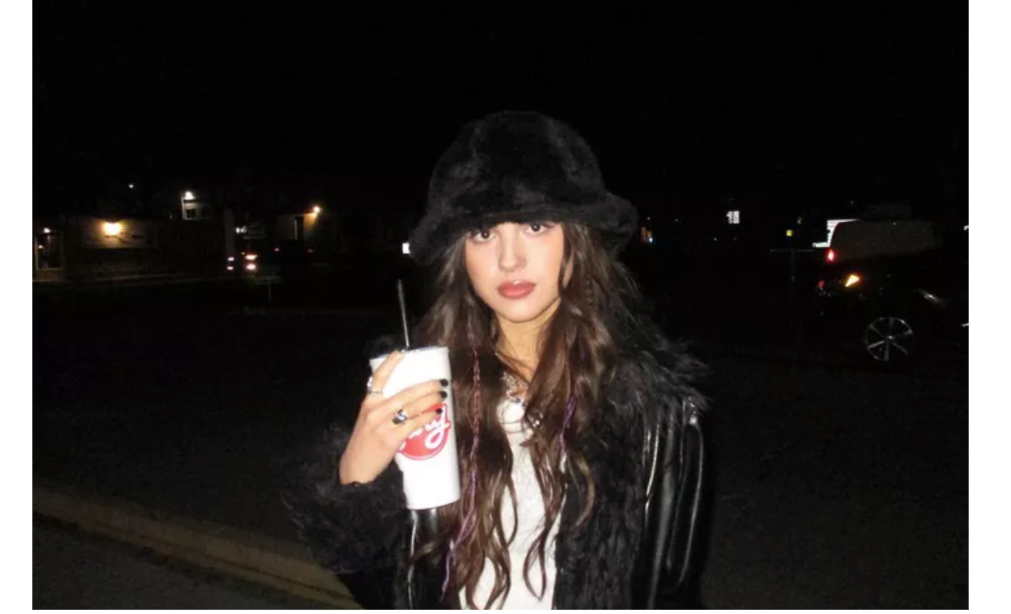 Olivia Rodrigo stands in black leather jacket and black hat with a large Swig soda. 