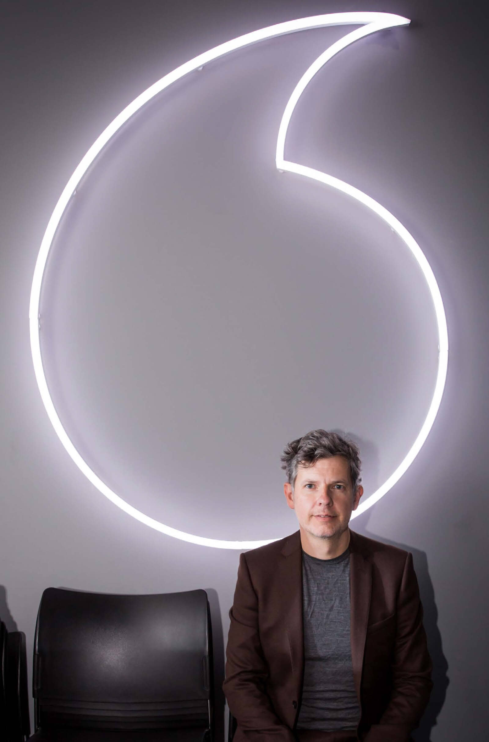 A man sits in front of a large neon quotation mark sign