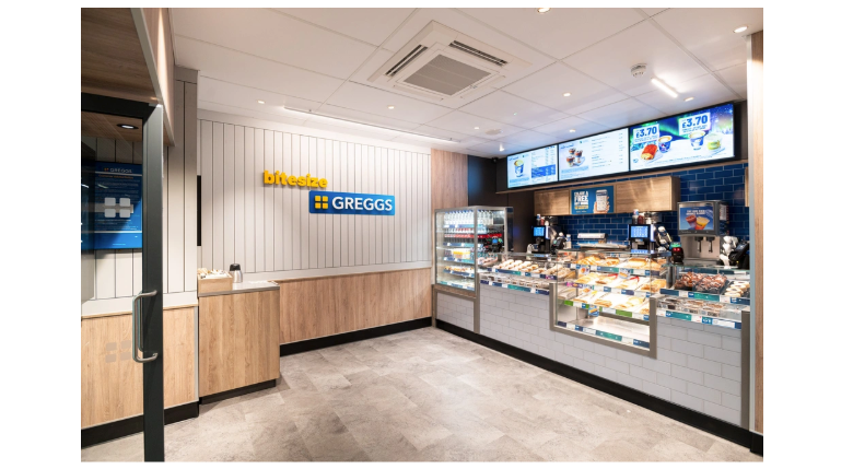 Photo of the interior of a small-format Greggs, with a counter and display case along one wall, and the brand’s logo on another.