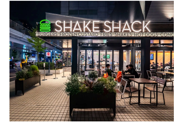 Photo of the outside of a Shake Shack location, with outdoor seating, a neon sign with a burger outlined in green, and the words “Shake Shack.”