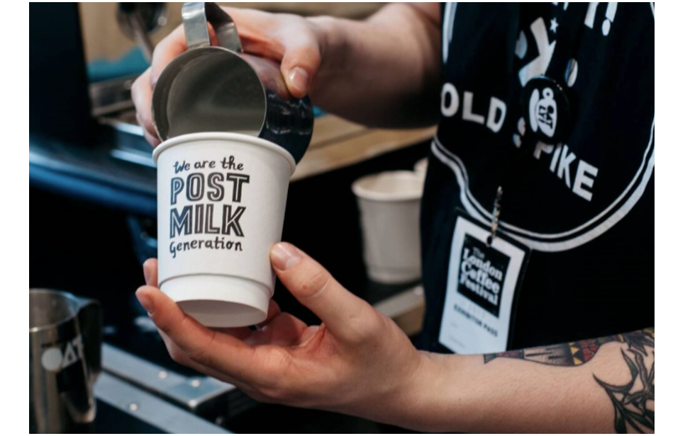 Oatly’s paper cup with the wording, “We are the post milk generation,” is being poured into by a barista with tattoos and a lanyard that readers, “The London Coffee Festival,” around their neck. 