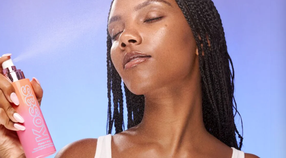 Woman sprays her face with a bottle of Kosas setting spray. 