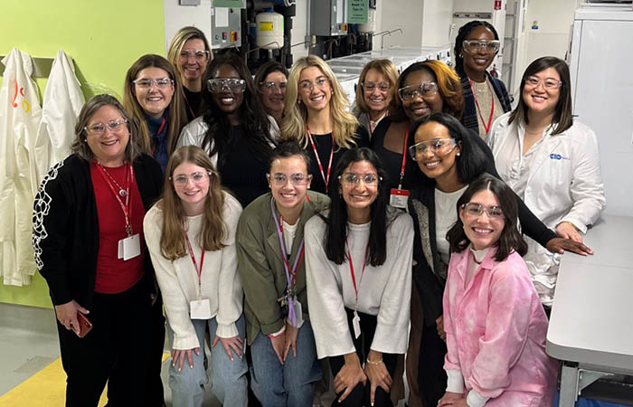 A large group of women and girls pose together and smile. They are standing in a laboratory and wearing protective eye gear.