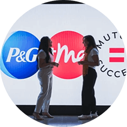 Two women face each other as they engage in conversation. They are standing at the end of a hallway, in front of a digital display of a blue, white and pink logo.