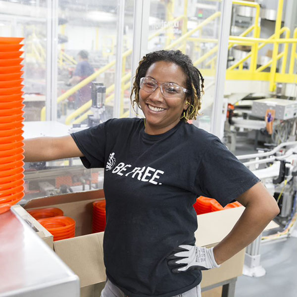A Black woman with dark curly hair. She stands inside a manufacturing plant, with large machinery in the background.