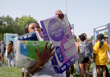 A young Black woman smiles as she carries an armful of household products, such as paper towels and toilet paper.