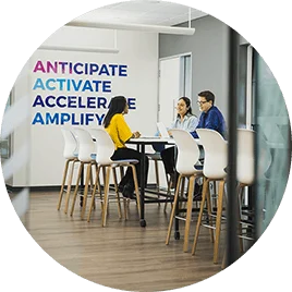 Three office employees work together on their laptops at a high-top table, in front of a sign that says "Anticipate; Activate; Accelerate; Amplify."