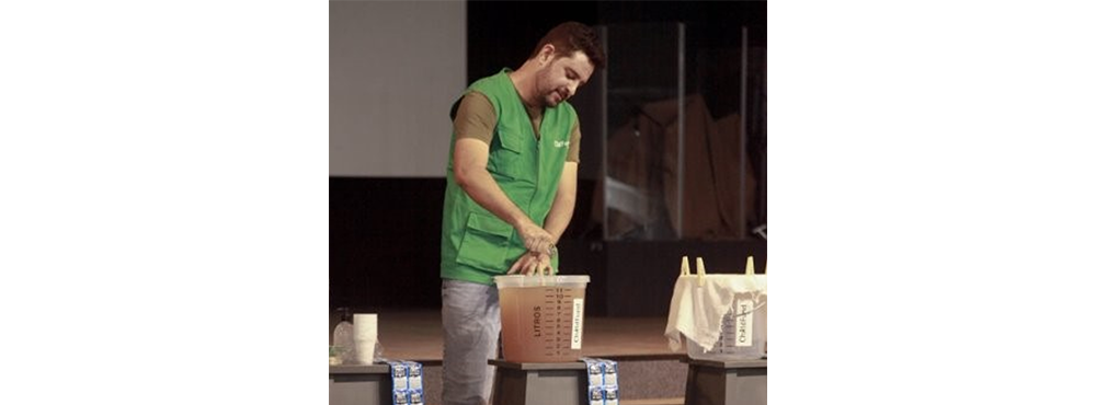 A man in a green vest stands in front of a transparent bucket of water.