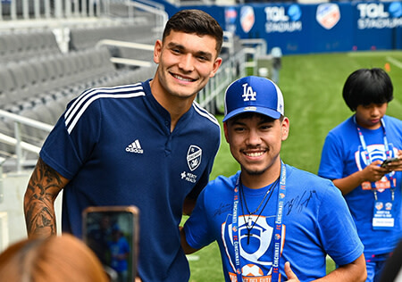 A Hispanic male in a dark blue soccer uniform. He stands next to a young Hispanic male student in a bright blue hat and t-shirt. They are standing on a soccer field.