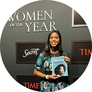 Smiling Black woman standing in front of a 'Women of the Year' PR wall and looking at the camera