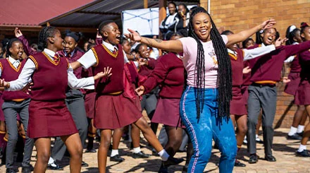 A Black woman with long, dark braided hair. She leads a larger group of female students in dance. The girls wear red and white uniforms.