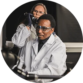 A man and woman in white lab coats. The woman holds up a tool to the man's head for a hair analysis.