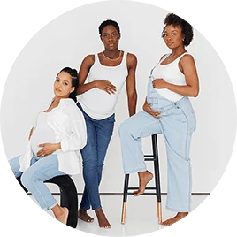 Three Black women in white shirts and blue jeans. All three women are pregnant and pose while standing or sitting on stools.