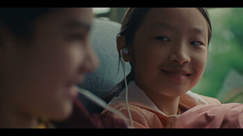 A young Asian girl on a school bus. She is smiling at another young girl who sits next to her, and they are sharing headphones.