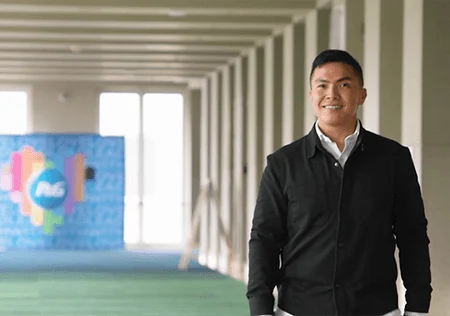 An Asian man stands in the hallways of his office and smiles at the camera.