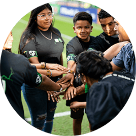 Several young male and female Hispanic children. They are standing in a circle on a soccer field and joining hands in the middle.