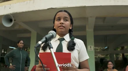 A young Indian girl in a white and green school uniform. She stands at a microphone while holding a red piece of paper.