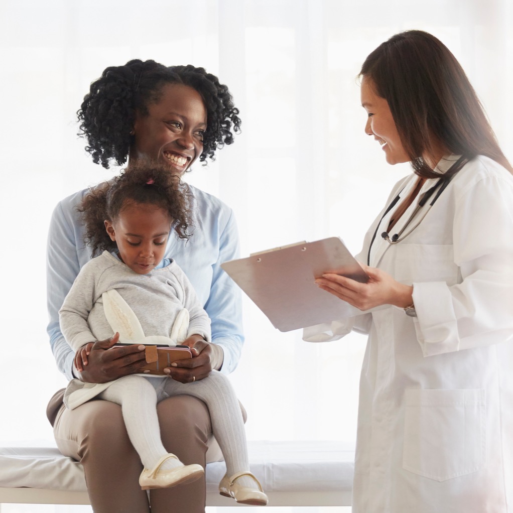 A doctor talking to a mother and child. The child looks at a phone while the mother and doctor smile.