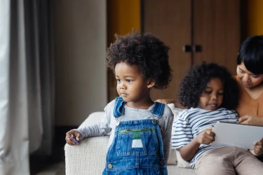 A child in overalls leans against a couch and looks off camera, while another child sits beside him looking at a tablet.