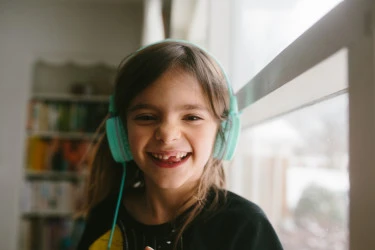A girl with a wide grin and missing front tooth wearing teal over-ear headphones and looking at the camera