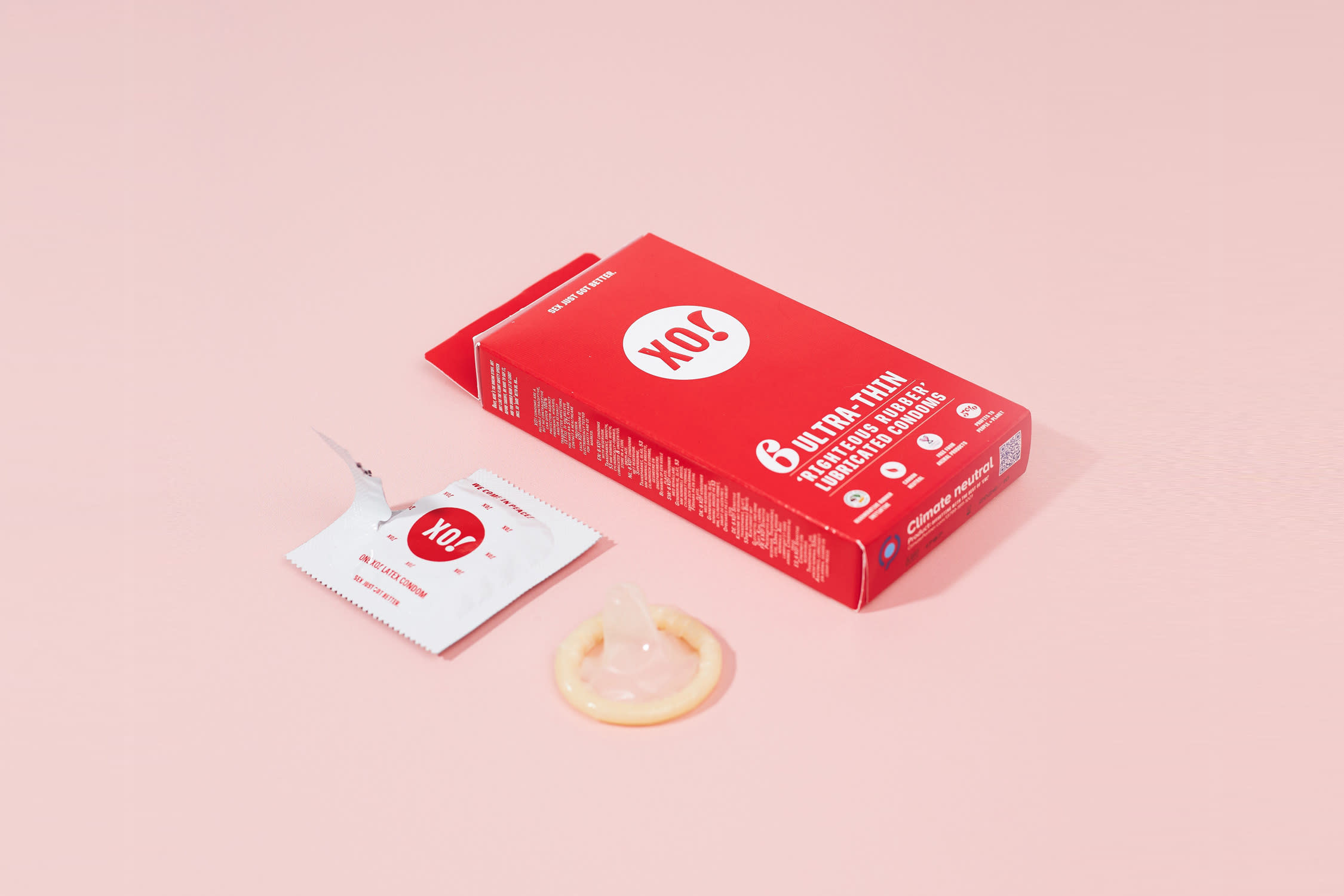 A red box of condoms on a pale pink background. One condom has been removed from the pack and taken out of the foil wrapper. The wrapper and condom sit next to the box.