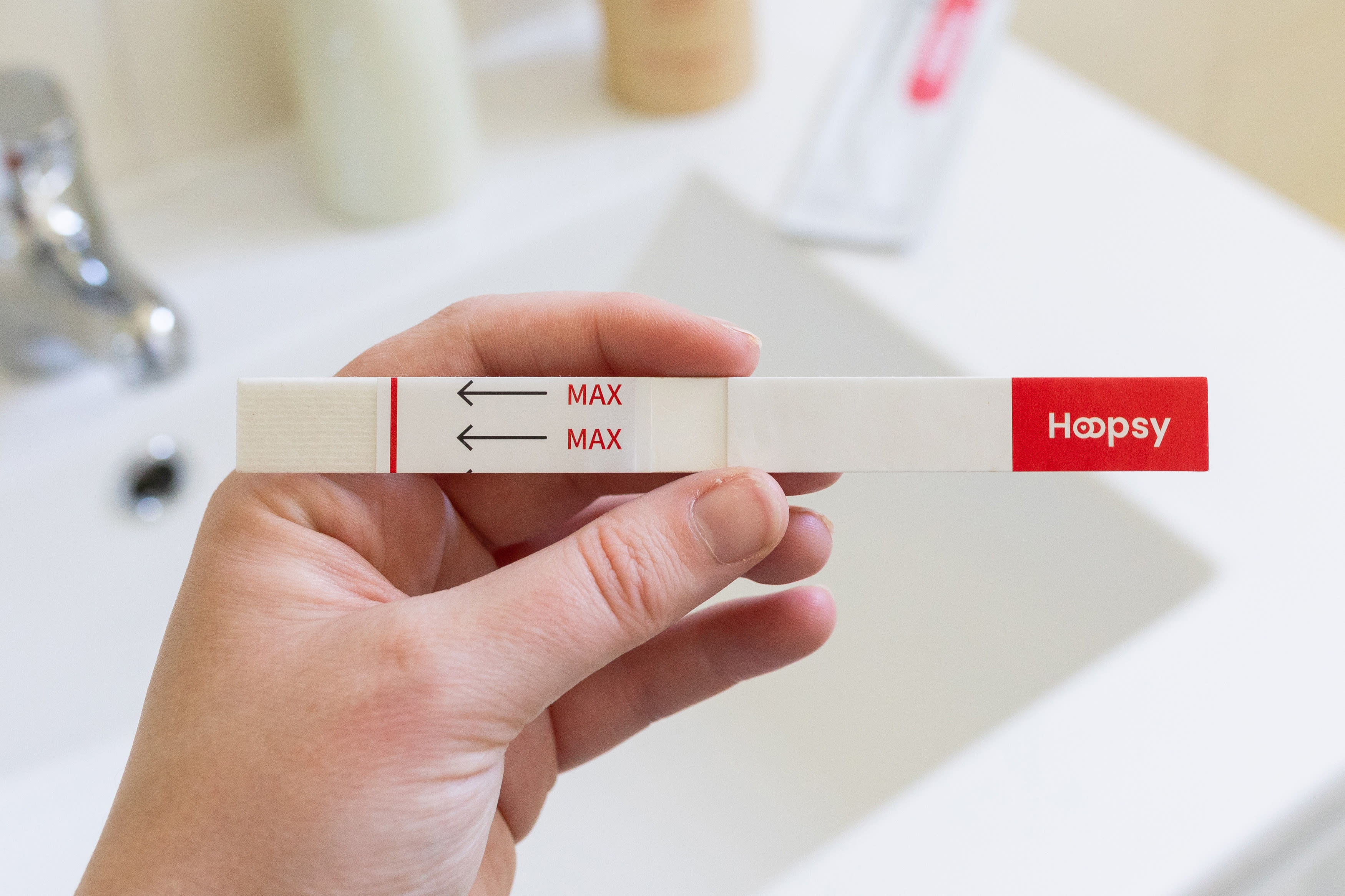 A white hand holding a pregnancy test strip over a sink. The test strip has a red Hoopsy logo at one end.